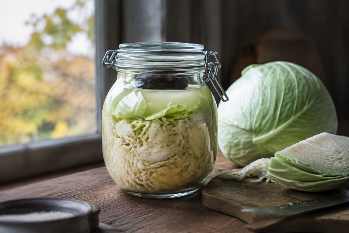 Holzgabel hebt Sauerkraut aus einem mit Lake gefüllten Bügelglas neben Weißkohl und Salz.